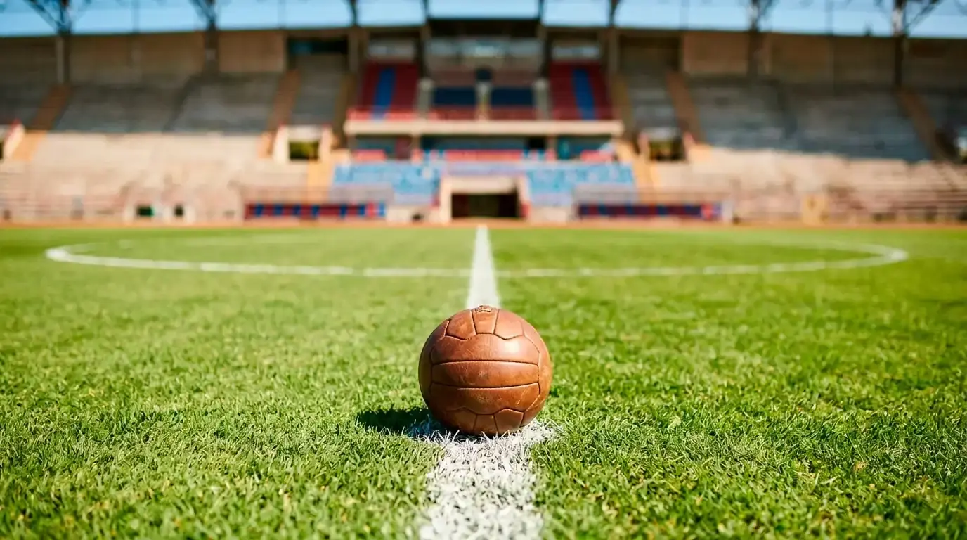 A football resting on the halfway line of a sunlit stadium pitch with empty stands behind