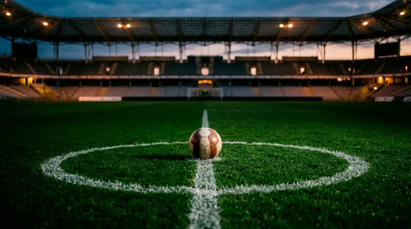 A football resting on the centre circle of an empty floodlit stadium pitch at dusk