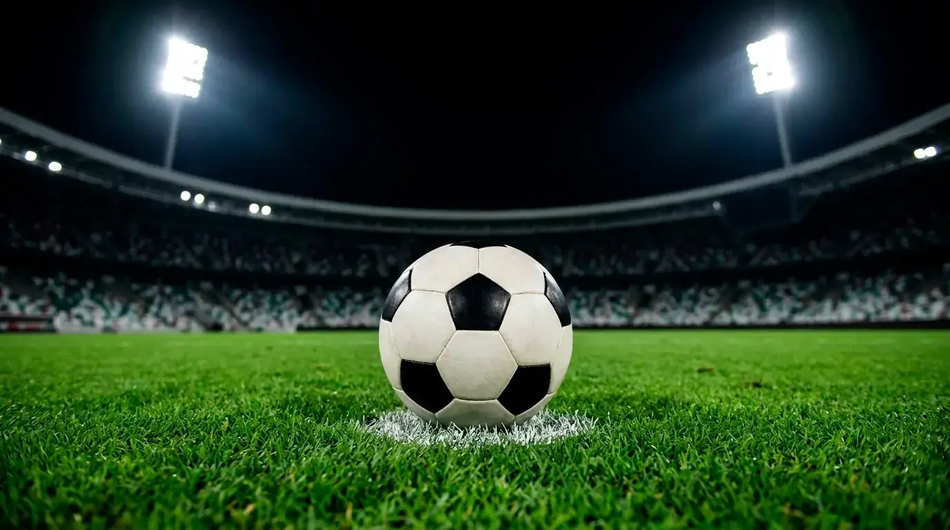 A football resting on the penalty spot of a floodlit pitch at night, empty stands behind
