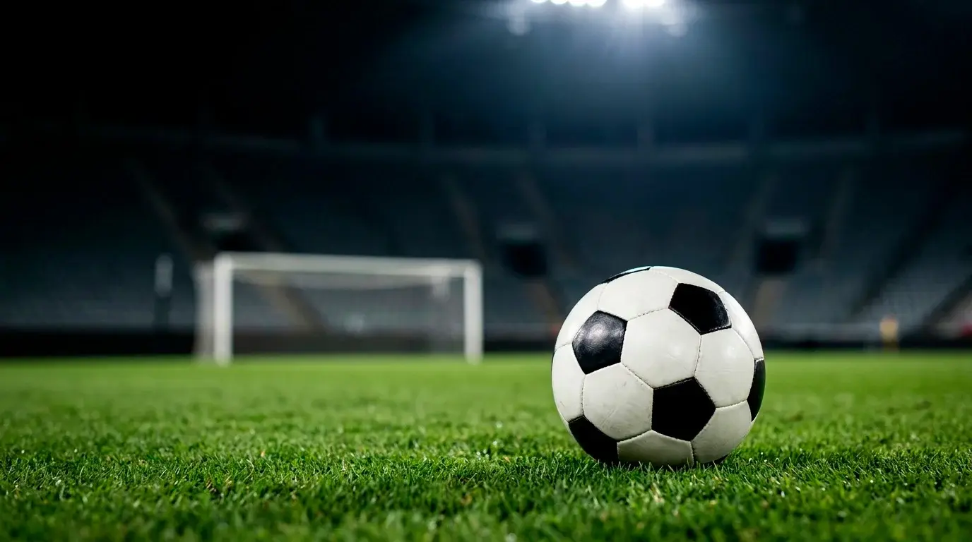 A close-up of a football on a floodlit pitch at night with empty stands behind