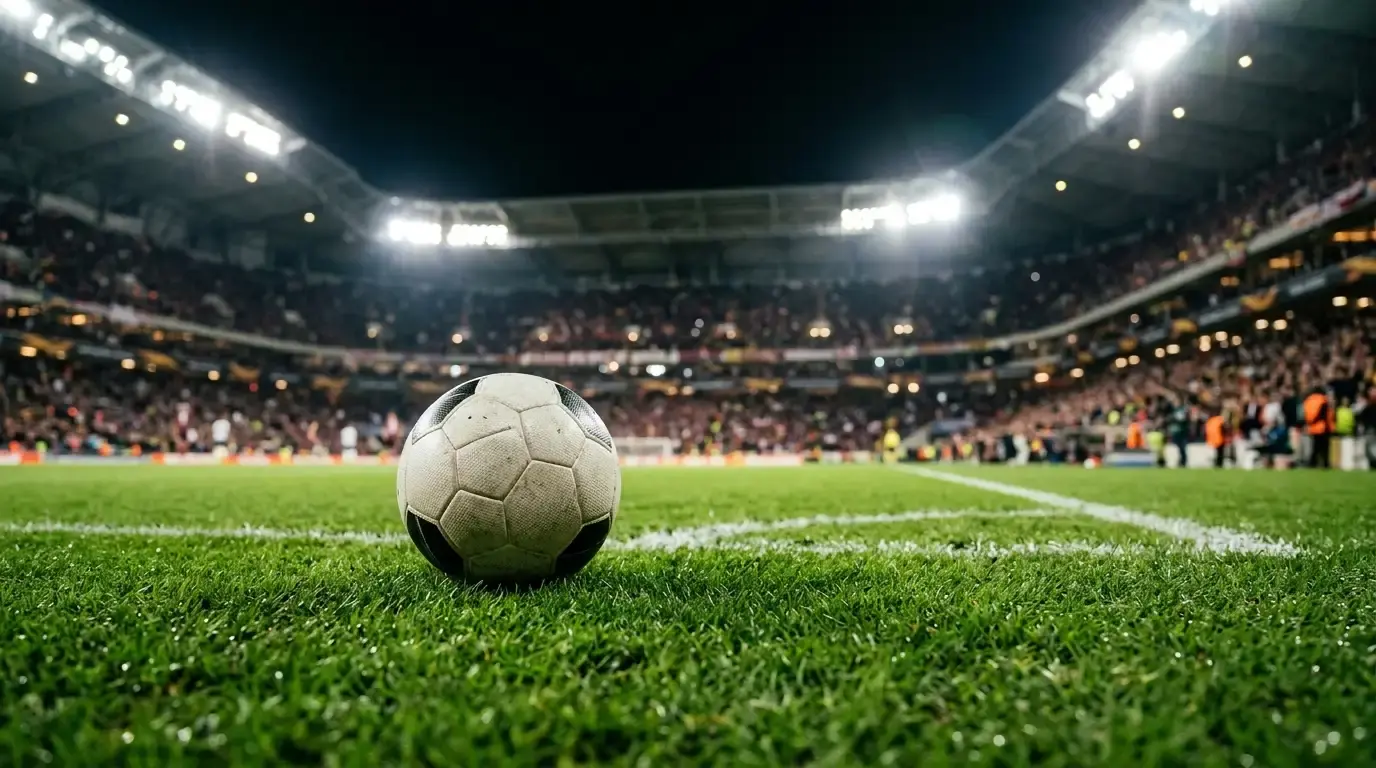 A close-up of a football on a floodlit pitch under bright stadium lights, the stands a soft blur behind